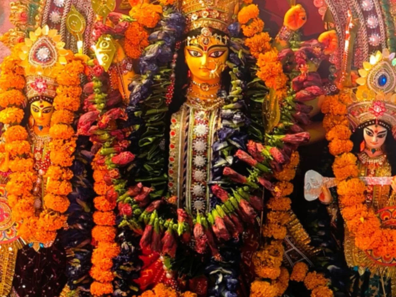 A vibrant, close-up shot of the traditionally decorated Durga idol at Jagriti Dham, flanked by Goddesses Lakshmi and Saraswati, all adorned with heavy garlands of marigold flowers and intricate jewelry.
