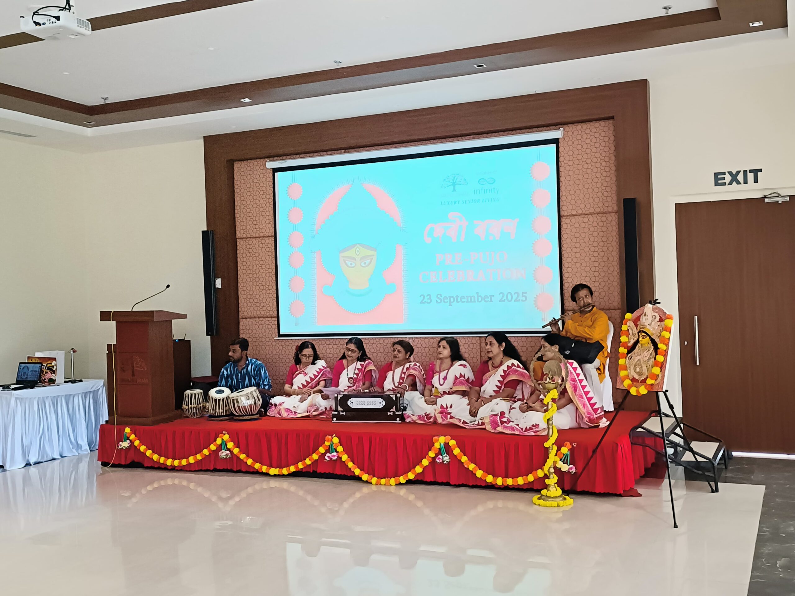 Musicians and singers from Team Sajhbela are on stage for a cultural programme at Jagriti Dham. Women in beautiful matching sarees sing into microphones, accompanied by men playing the tabla and flute, as part of the Pre-Puja celebration at Jagriti Dham.