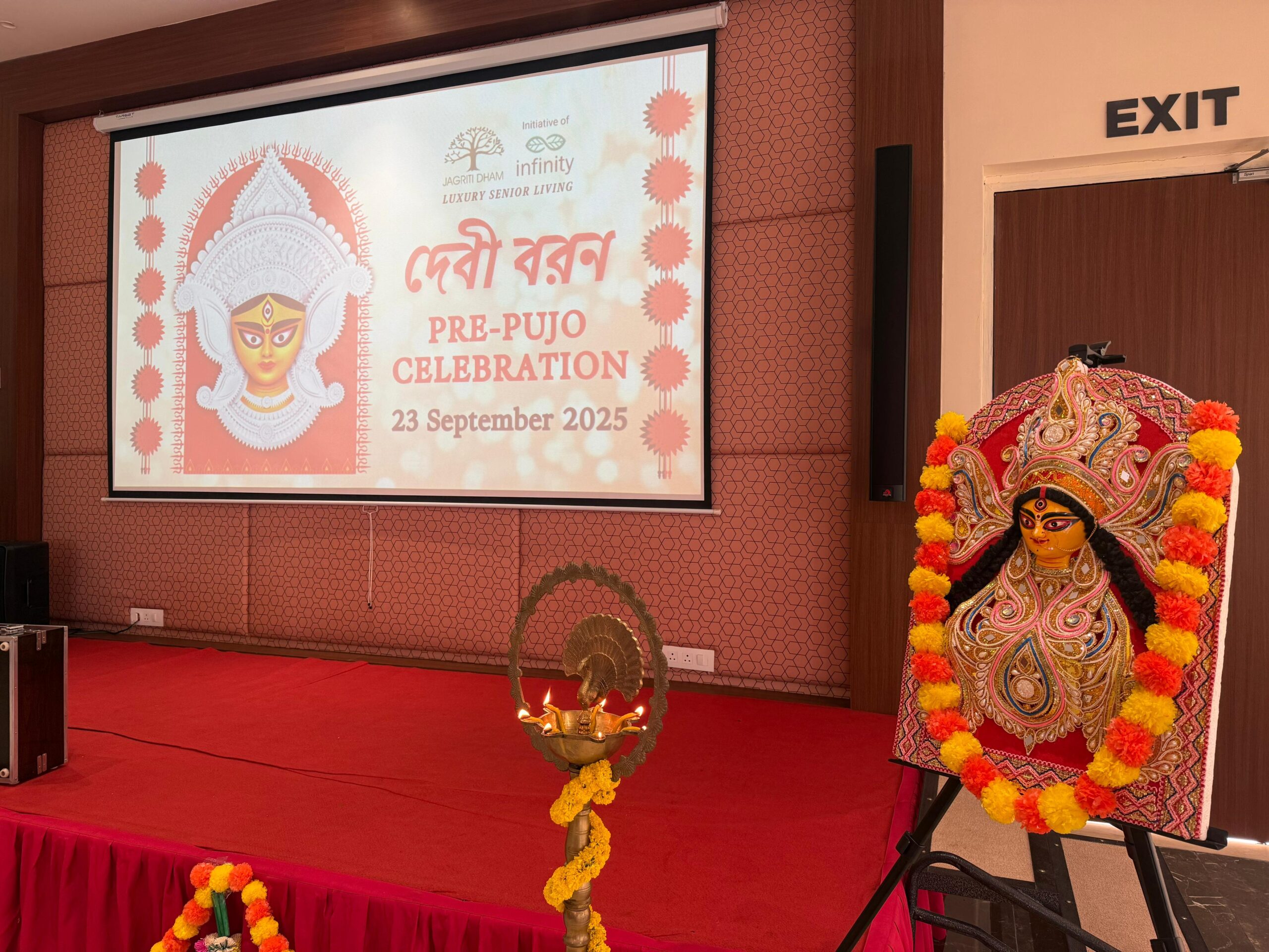 A decorated stage at Jagriti Dham set for a 'Debi Boron' Pre-Pujo Celebration, featuring a festive screen, a lit traditional lamp, and an idol of Goddess Durga.