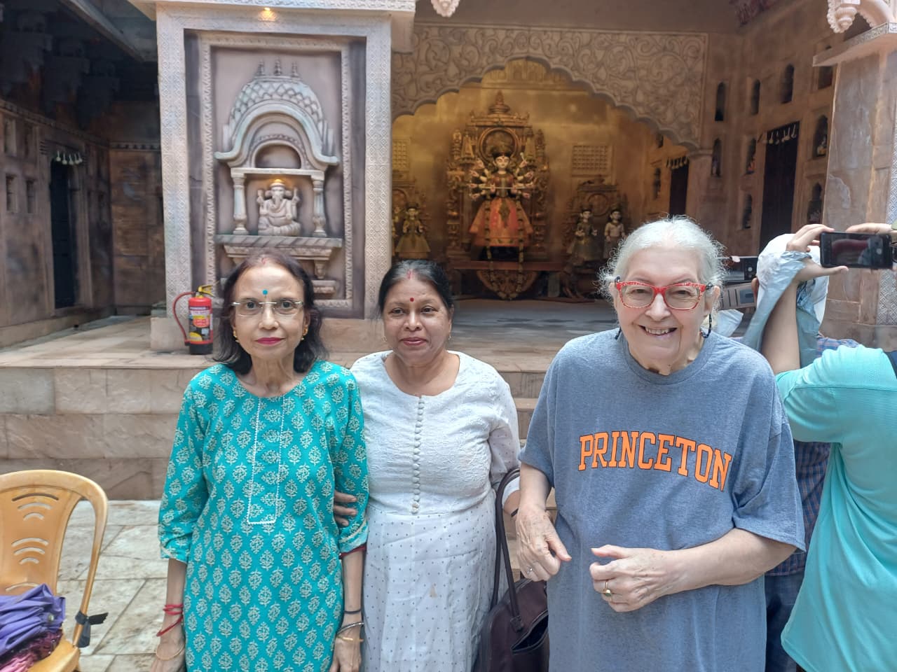 Three smiling senior residents from Jagriti Dham enjoying a pandal hopping trip on Sasthi, posing together in front of a beautiful Durga idol.