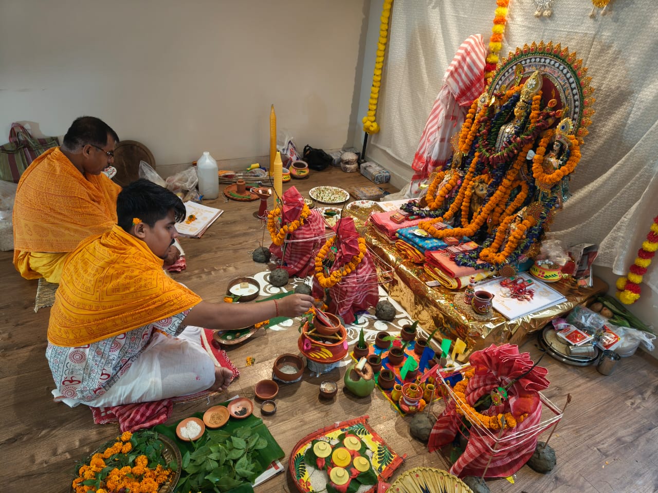 Two priests in traditional orange and white attire perform sacred rituals for Durga Puja at Jagriti Dham, seated on the floor before the idol with various ceremonial items and decorated pots (ghots).