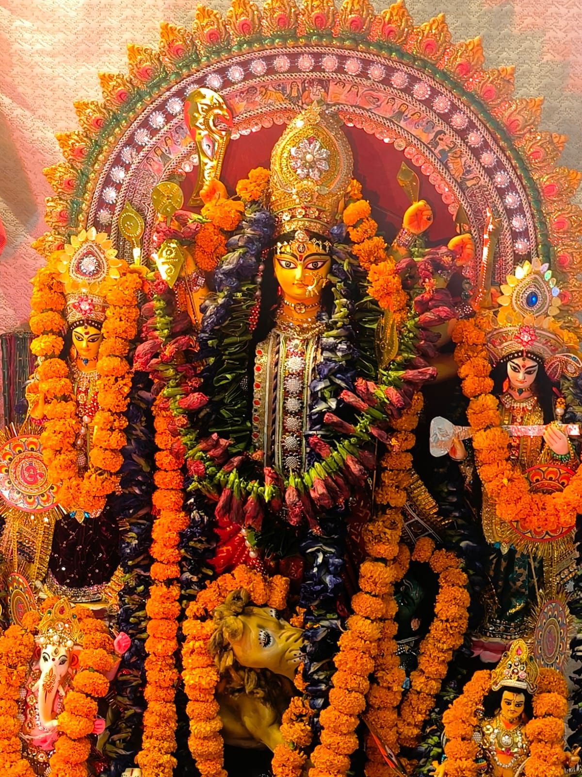 A vibrant, close-up shot of the traditionally decorated Durga idol at Jagriti Dham, flanked by Goddesses Lakshmi and Saraswati, all adorned with heavy garlands of marigold flowers and intricate jewelry.
