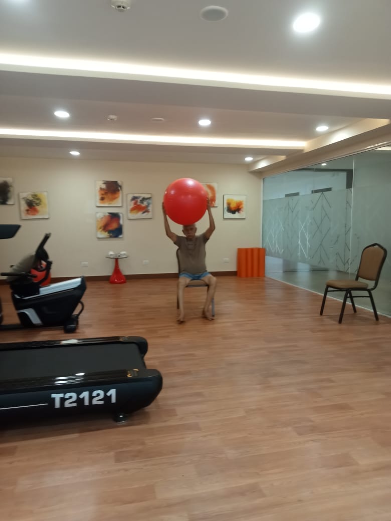 A senior resident performing seated strength training exercises with a gym ball in the modern fitness centre of a premium Assisted Living facility in Kolkata.