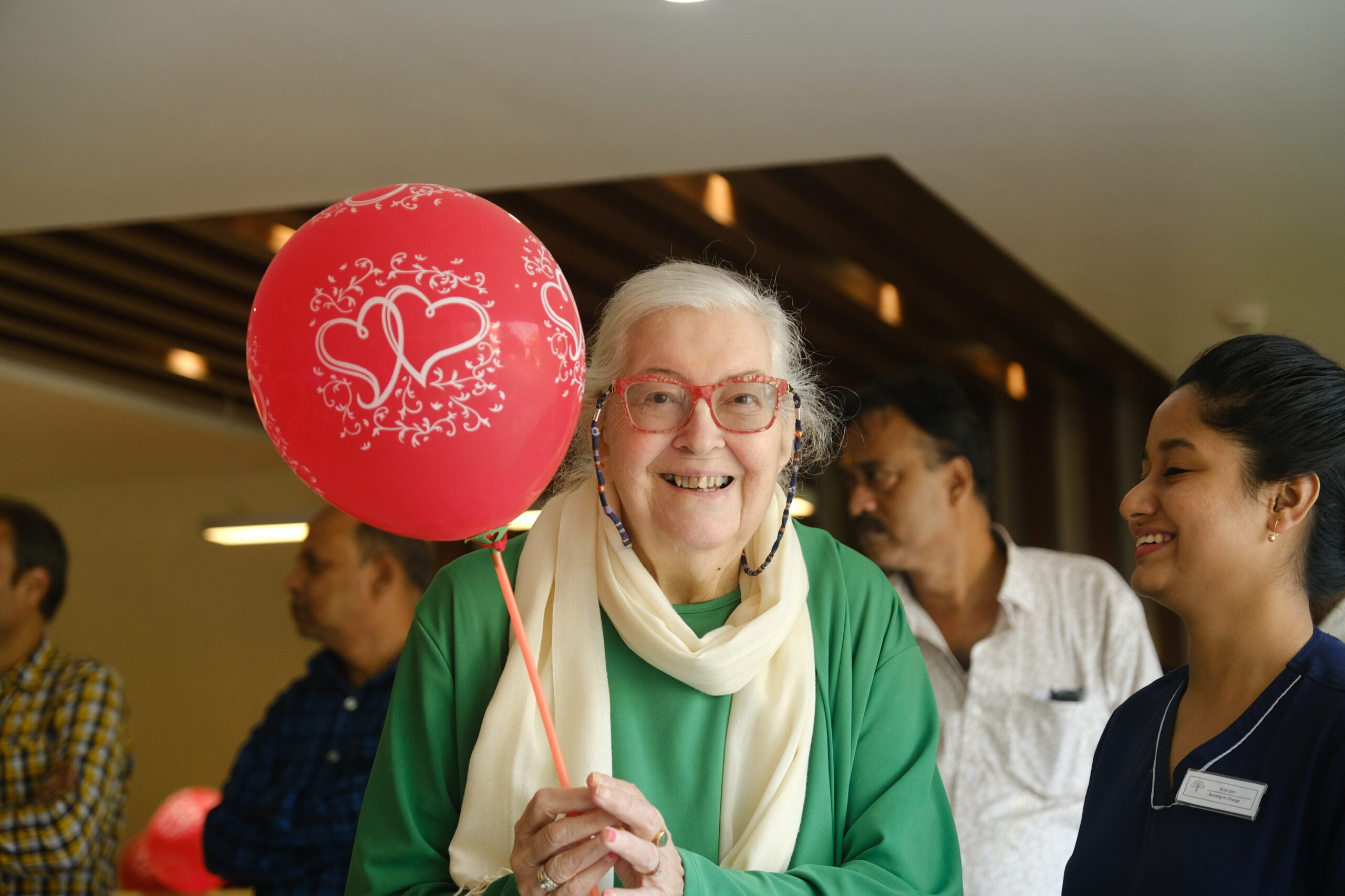 Jagriti Dham resident Greeta Aunty smiling joyfully while holding a red balloon during the Metta Foundation's "Celebrating Life Across Generations" event.