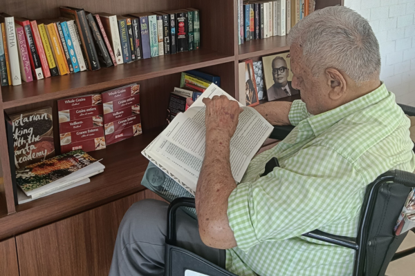 An elderly male resident sitting in a wheelchair, reading a book intently in front of a well-stocked wooden bookshelf at the Jagriti Dham library.
