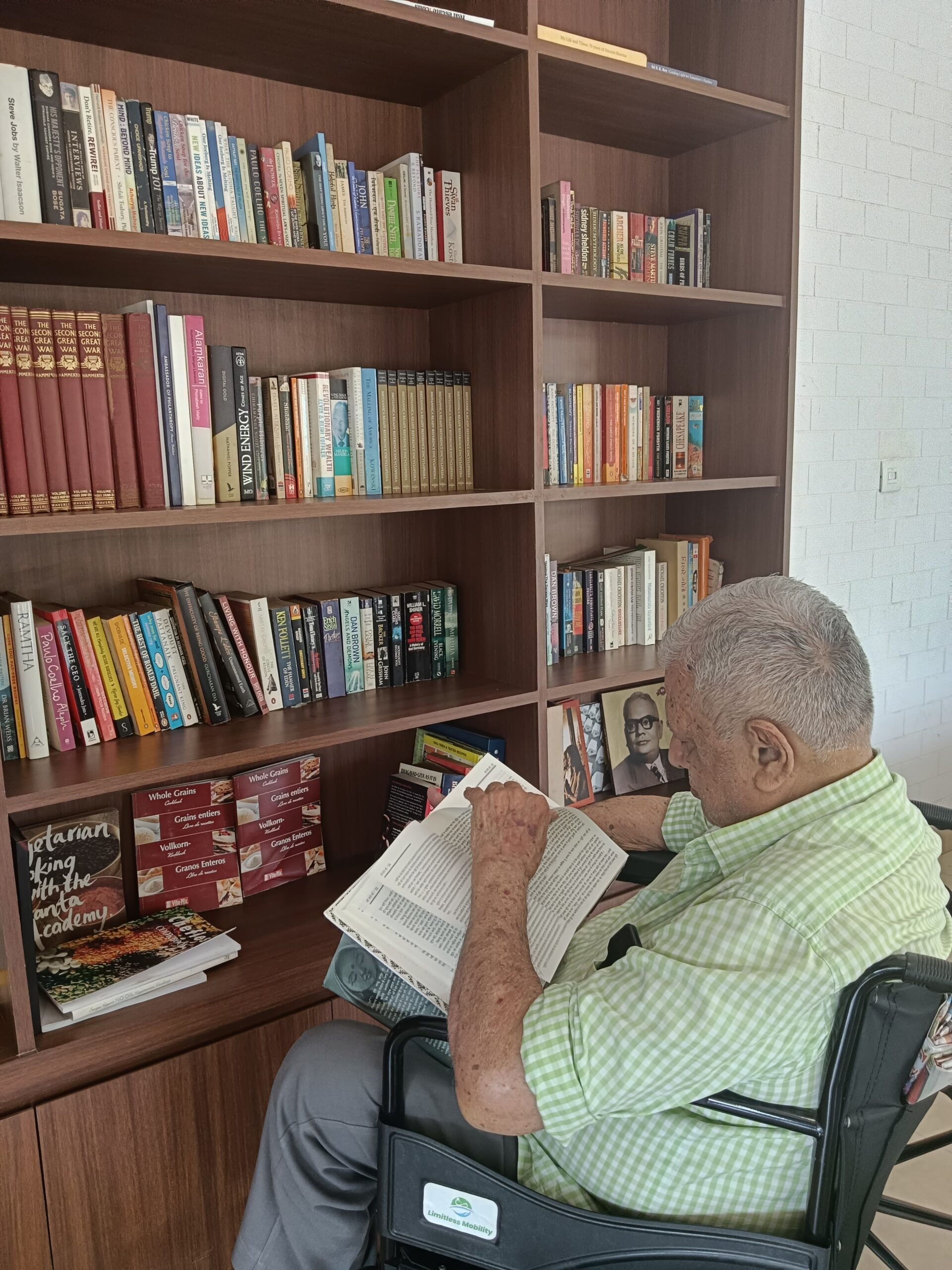 An elderly male resident sitting in a wheelchair, reading a book intently in front of a well-stocked wooden bookshelf at the Jagriti Dham library.