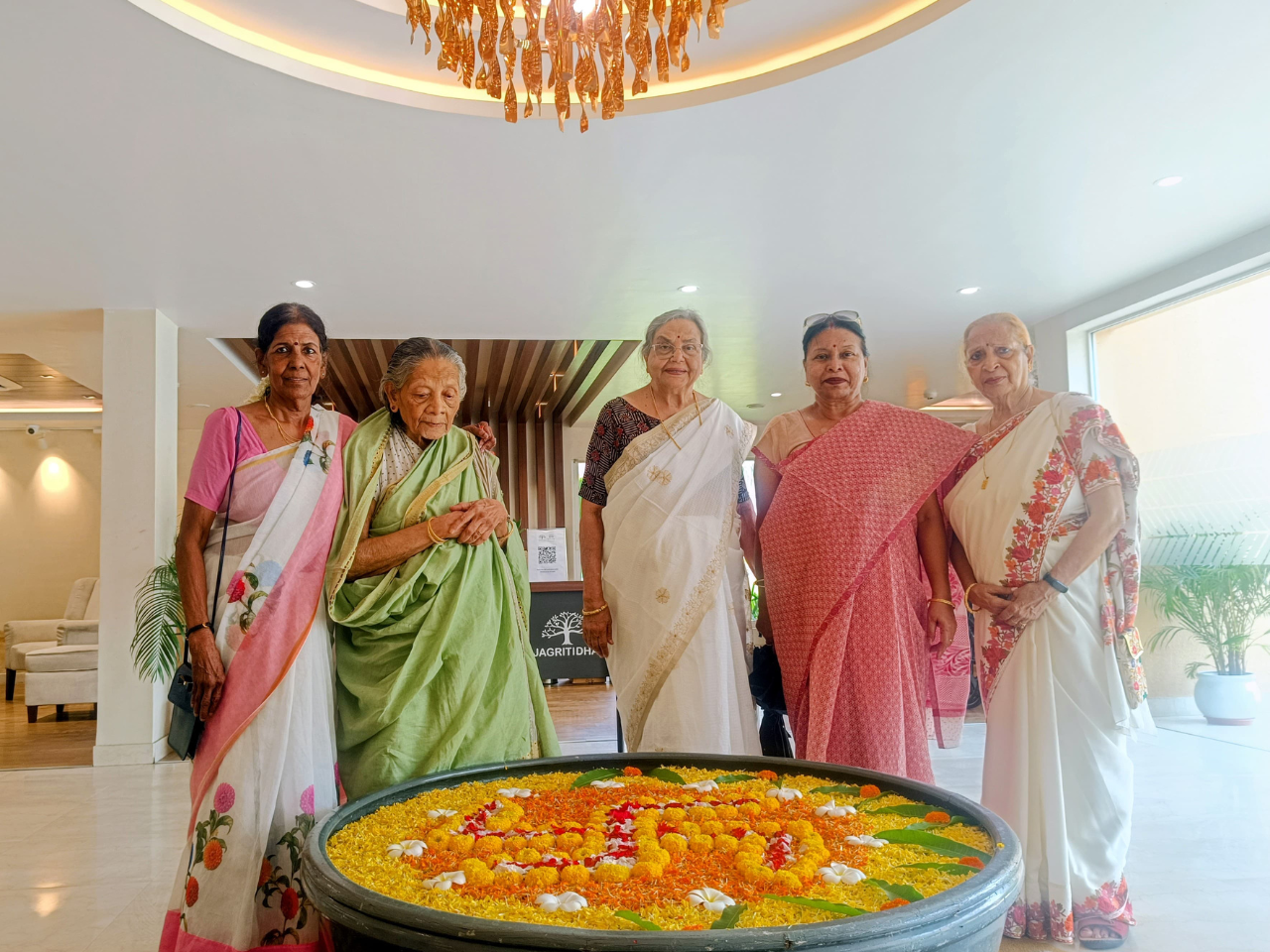 Five senior women in traditional Bengali sarees gather around a large bowl of water decorated with marigolds in the modern lobby of Jagriti dham a luxurious old age home in Kolkata, with natural light pouring in.