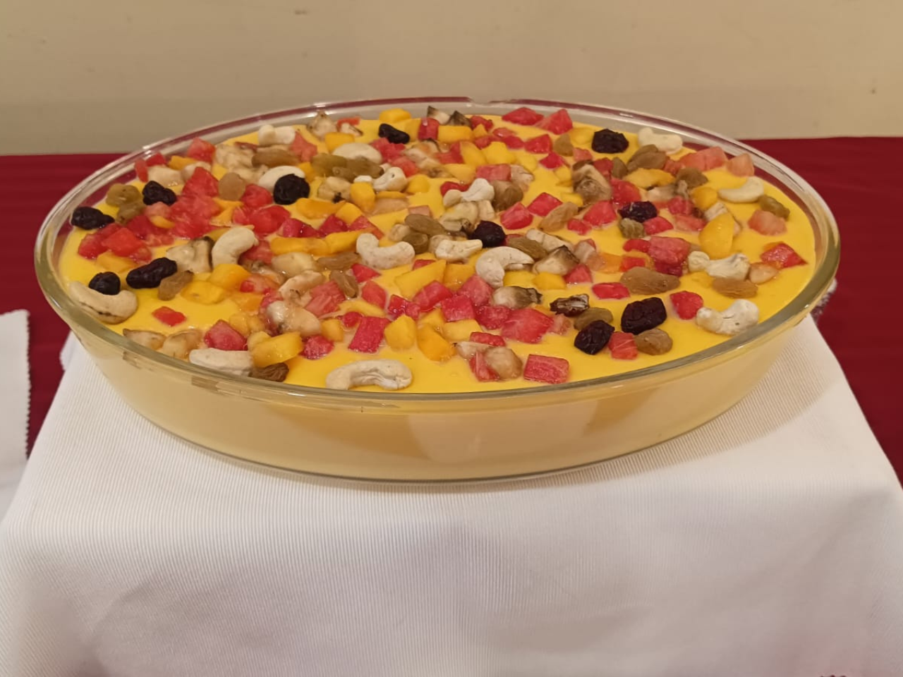 A large, appetizing bowl of creamy fruit custard dessert, generously topped with fresh diced fruits, raisins, and cashew nuts prepared specially for the senior residents at Jagriti Dham.