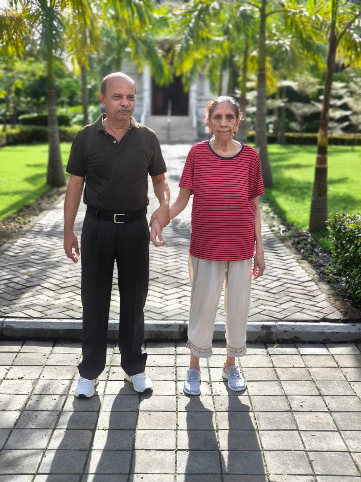 An resident holding hands with a caregiver on a paved pathway near the Krishna Temple at Jagriti Dham, a premium facility providing housing for senior citizens in a lush, green environment.