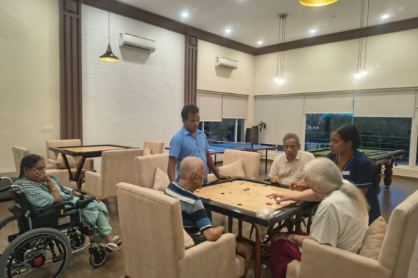 Senior residents engaging in a game of carrom in the spacious indoor games room of Jagriti Dham, a luxurious old age home in Kolkata, with staff members providing support and companionship.