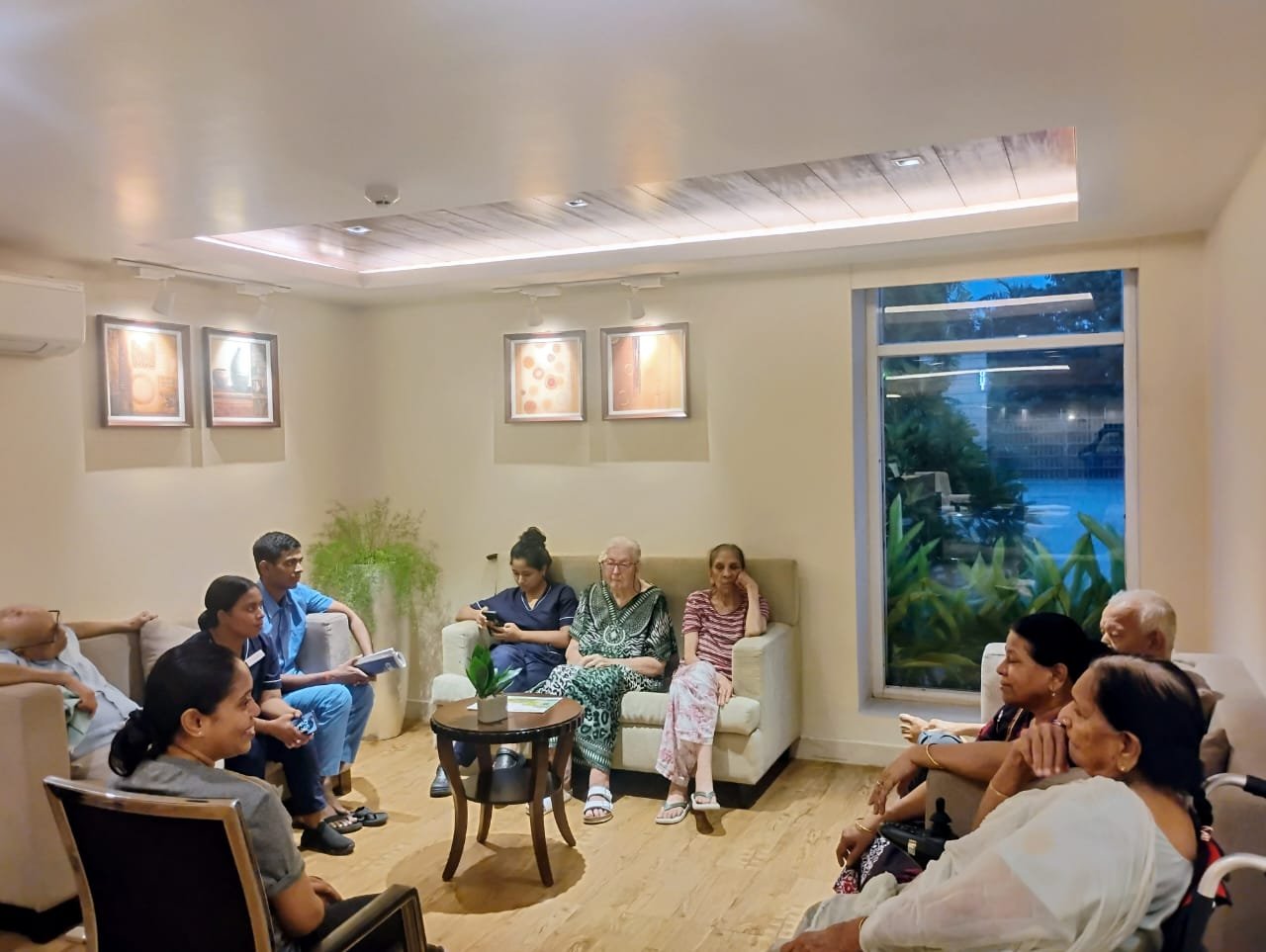 A group of senior residents and young caregivers are gathered in a modern, warm, and comfortable lounge area at Jagriti Dham, engaging in an "adda" (informal conversation) session, as seen in this photo from one of the finest luxury senior living facilities. Diverse seniors are seated on plush sofas and chairs, all facing each other and interacting with a few staff members in scrubs. The room is decorated with artwork, a central coffee table, recessed lighting in a wood-and-tray ceiling, and a large window in the background looking out onto a dark, lush garden, creating a peaceful and high-end environment for social connection.