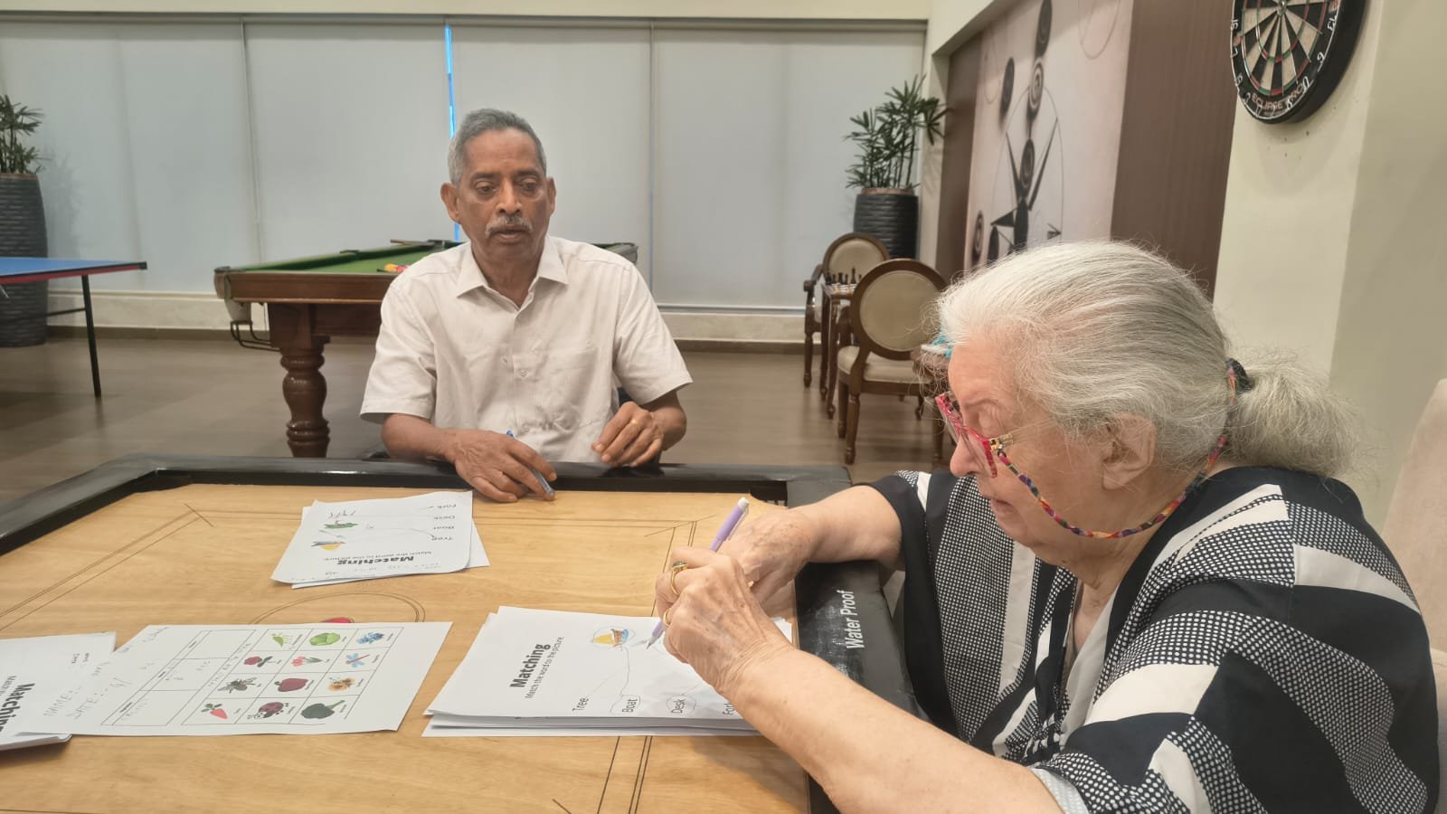 Two elderly residents participating in a cognitive session, working on matching worksheets at a table in the games room of Jagriti Dham, highlighting the engaging mental wellness activities offered at senior living communities in India.