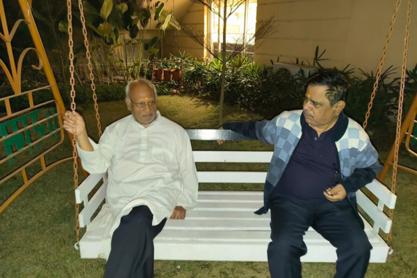 Two senior male residents of Jagriti Dham, a senior living community, are sitting together on a white wooden bench swing on a well-lit garden lawn at night, spending evening time together and engaged in conversation. The older man on the left, with white hair and a beard, wears a white kurta-pajama and has a tilak on his forehead. He is holding a swing chain and facing the other man. The second man, in a blue and white patterned sweater over a dark shirt and also with a tilak, listens and speaks. They are on a lawn with green plants and a building wall with a lit window visible in the background. The swing is supported by a yellow-painted metal frame.