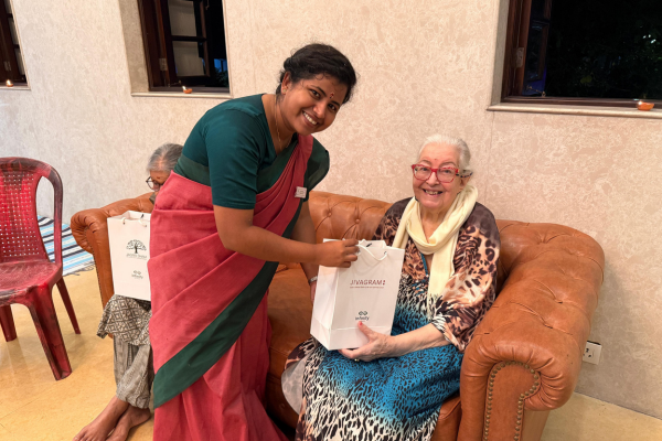 A smiling staff member in a traditional saree hands a Diwali gift bag to a cheerful elderly resident sitting on a sofa at Jagriti Dham, recognized among the best old age homes in Kolkata.