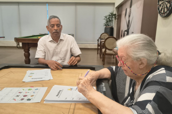 Two elderly residents participating in a cognitive session, working on matching worksheets at a table in the games room of Jagriti Dham, highlighting the engaging mental wellness activities offered at senior living communities in India.