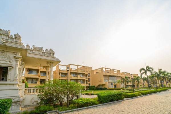 Scenic exterior view of a premium residential complex, representing Jagriti Dham - one of the best old age homes in Kolkata, featuring multi-story buildings, lush landscaped gardens, palm trees, and a peaceful paved walkway.
