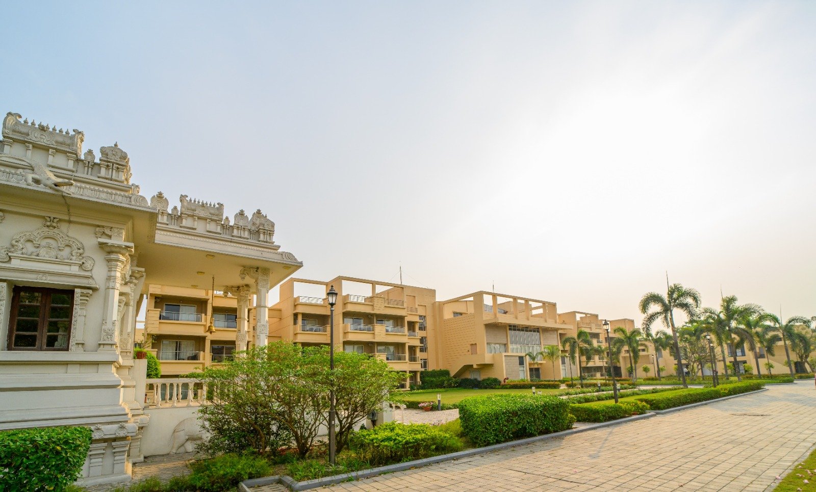 Scenic exterior view of a premium residential complex, representing Jagriti Dham - one of the best old age homes in Kolkata, featuring multi-story buildings, lush landscaped gardens, palm trees, and a peaceful paved walkway.