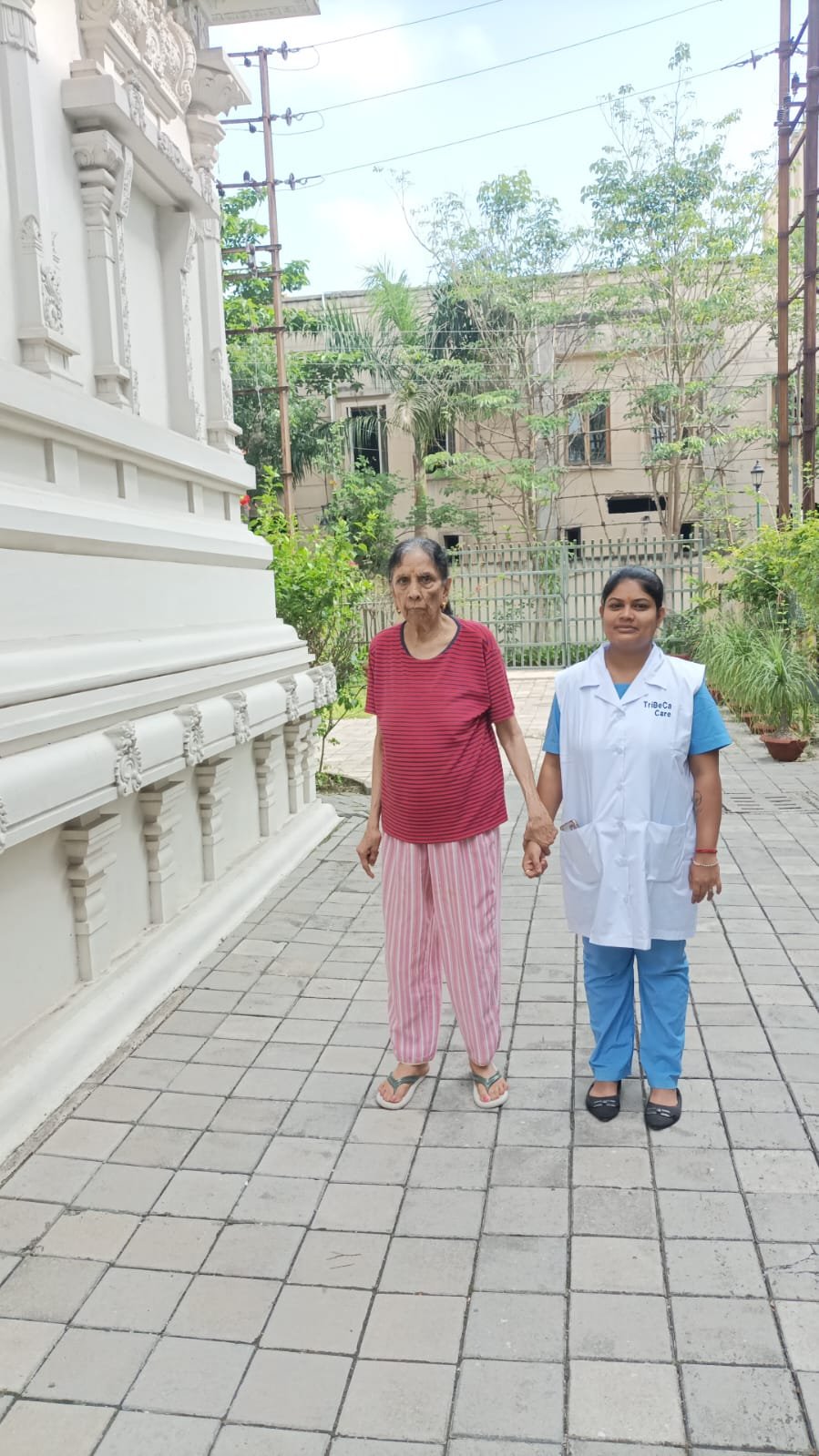 A full-length view of an elderly female resident wearing a red striped top and pink striped trousers, holding hands with a smiling caregiver dressed in blue scrubs and a white 'TriBeCa Care' coat. They are standing on a grey stone-paved pathway beside a grand, ornately carved white building within the peaceful, greenery-filled campus of Jagriti dham - best old age homes in Kolkata.
