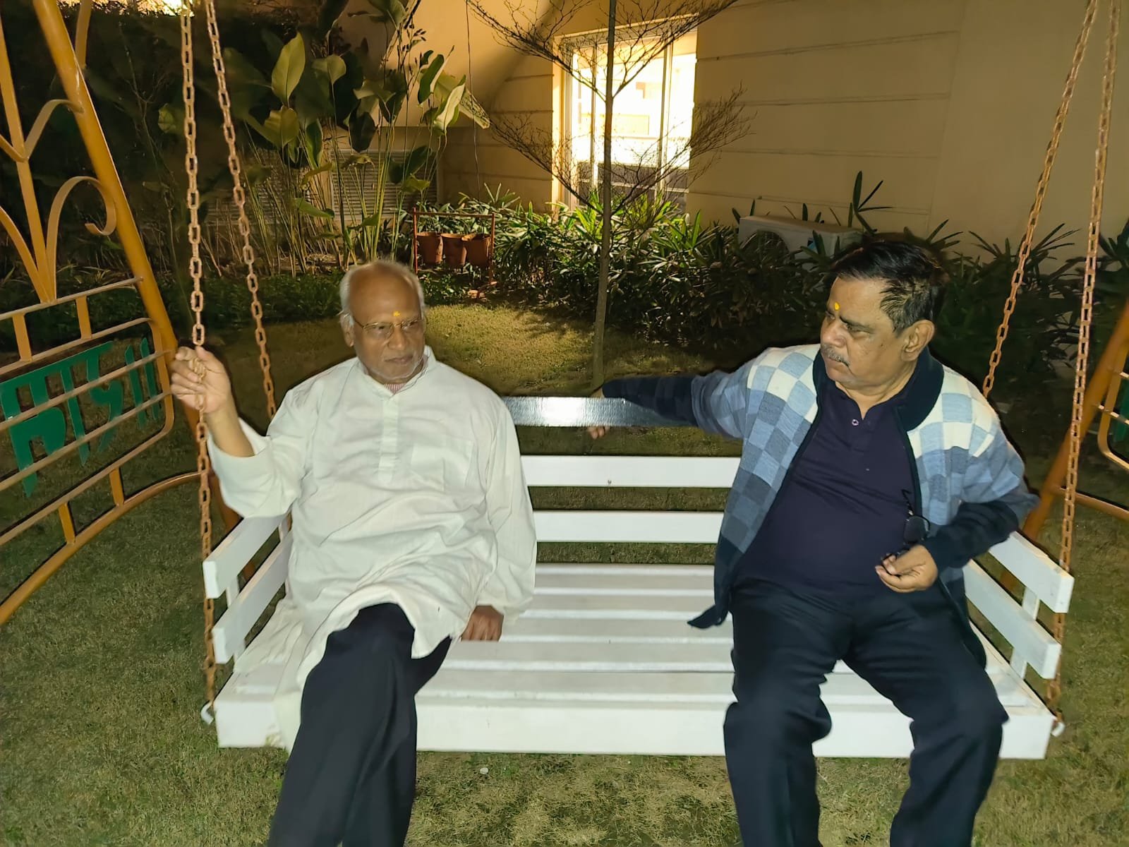 Two senior male residents of Jagriti Dham, a senior living community, are sitting together on a white wooden bench swing on a well-lit garden lawn at night, spending evening time together and engaged in conversation. The older man on the left, with white hair and a beard, wears a white kurta-pajama and has a tilak on his forehead. He is holding a swing chain and facing the other man. The second man, in a blue and white patterned sweater over a dark shirt and also with a tilak, listens and speaks. They are on a lawn with green plants and a building wall with a lit window visible in the background. The swing is supported by a yellow-painted metal frame.