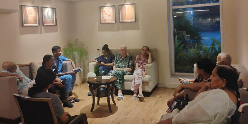 Group of senior residents in comfortable living room seating, including a man in patterned shorts, a woman in a green caftan, and others in casual wear and wheelchairs, gathered for a community presentation or meeting, with two male staff members in blue and black uniforms presenting from an armchair.