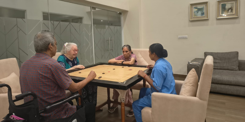 Four senior individuals, including three elderly residents and one caregiver in a blue uniform, gathered around a square carrom board playing a game in a well-lit indoor lounge of an old age home in South Kolkata