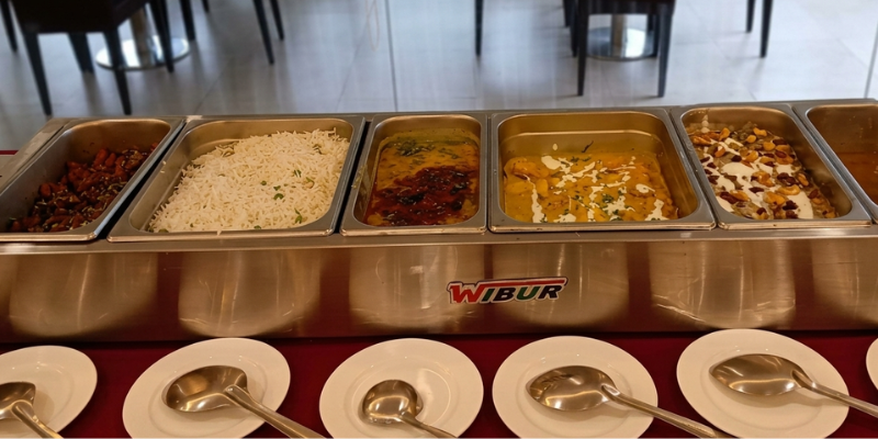 A clean buffet counter layout at the best old age home, displaying stainless steel chafing dishes filled with fresh Indian food items, including fried snacks, white rice, dal, rich curries, and a dessert topped with cream and nuts.