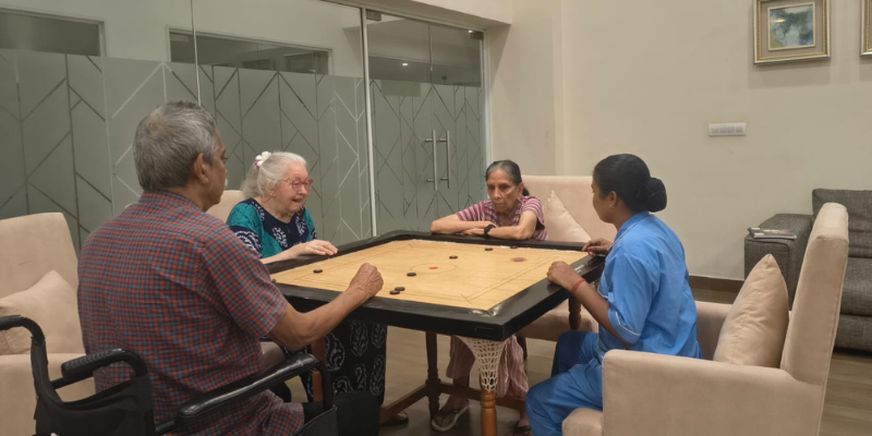 Senior residents enjoying a game of carrom with a dedicated caregiver in a modern indoor recreation room at a premium facility providing senior citizen care services.