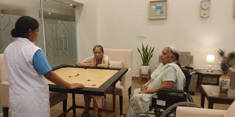 A group of people, including two senior residents and a facility staff member, are engaged in a game of carrom on a wooden tournament carrom board. One resident is in a wheelchair, and the staff member wears a white over-vest with a blue scrub-style top in a modern, comfortable living room with large glass doors, framed art, a large potted plant, and a table with peacock feathers.