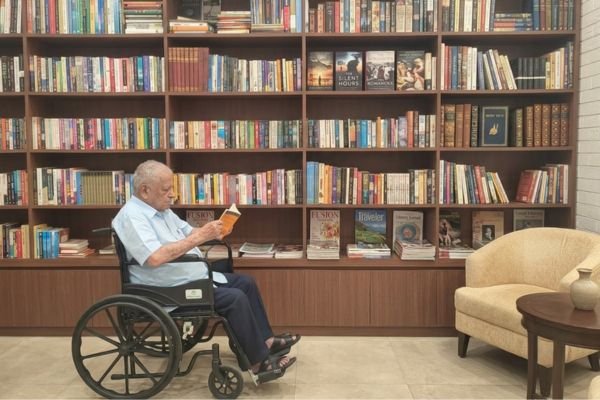 An elderly senior man sitting comfortably in a wheelchair, reading a book in a peaceful library lounge featuring an expansive, floor-to-ceiling wooden bookshelf filled with diverse books, alongside a cozy armchair and coffee table.