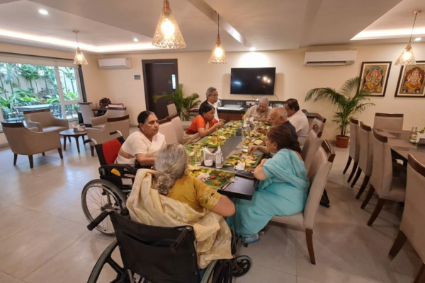 Senior residents enjoying a traditional Indian meal served on banana leaves inside a bright, modern dining room at a premium retirement facility, recognized as one of the best old age homes in Kolkata, with two residents sitting comfortably in wheelchairs at a long dining table.
