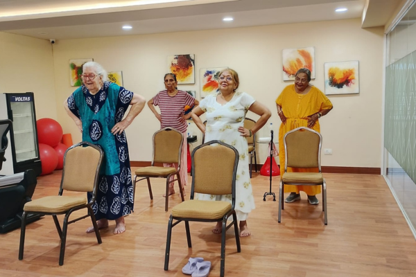 Four elderly senior women standing with their hands on their hips, participating in a gentle chair yoga or fitness class in a bright wellness studio room at a senior retirement community. The room features wooden floors, abstract artwork on the walls, individual padded chairs for support, and red exercise balls in the background.