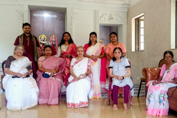 Senior citizens and family members gather for a group photograph during a community program celebrating older adults. Elderly women are seated in the front row, with younger family members standing behind them. Participants wear traditional sarees, and decorative idols in the background create a festive and culturally rich atmosphere.