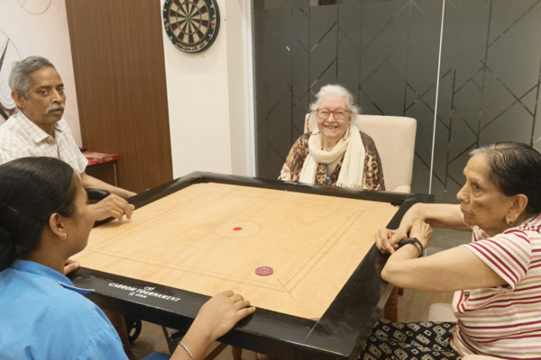 Four people, including senior residents and a caregiver in a light blue uniform, gathered around a wooden tournament carrom board playing a friendly match in an indoor recreational room at a senior living community.