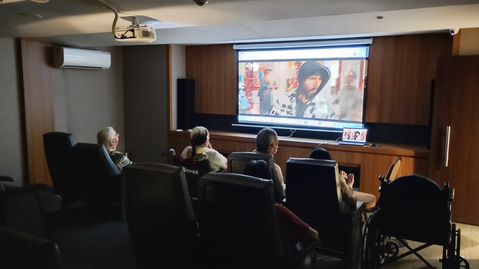 Senior residents enjoying a movie night in the luxury AV theatre at Jagriti Dham, a luxurious old age home seated comfortably in plush recliners and wheelchair-accessible spaces in front of a large projector screen.