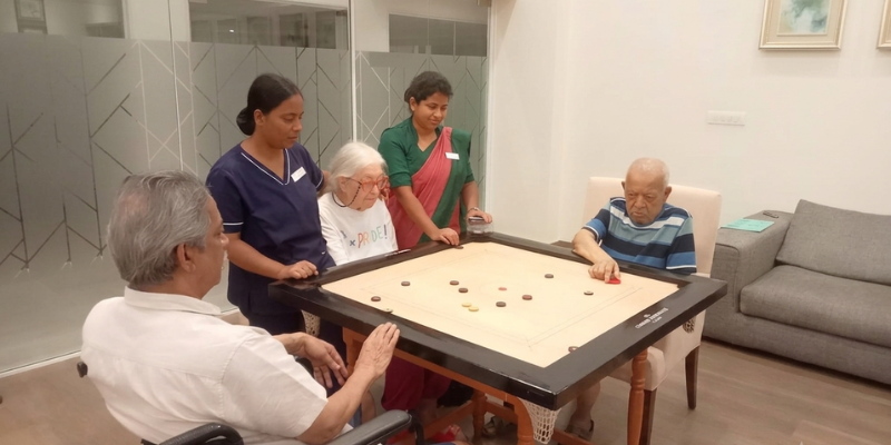 Senior residents participate in a carrom game with the support of caregivers in a comfortable indoor recreation area.