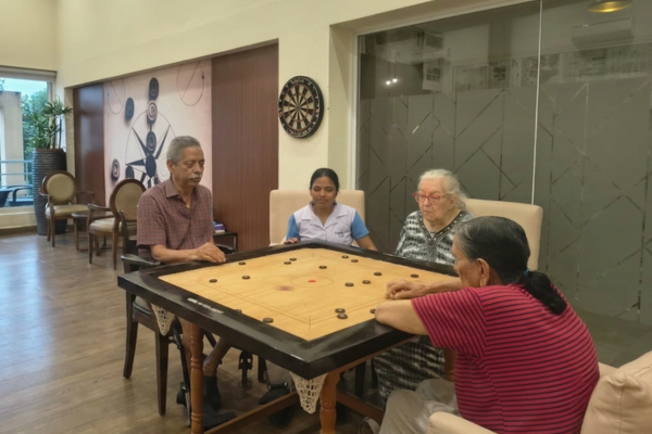A group of senior residents and an assistant gathered around a carrom board in an indoor games room, with the woman in the foreground wearing a pink outfit removed.