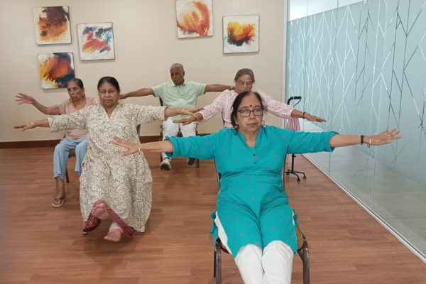 A group of senior citizens seated on chairs in a brightly lit indoor activity room, performing arm-extension exercises together during a wellness session.
