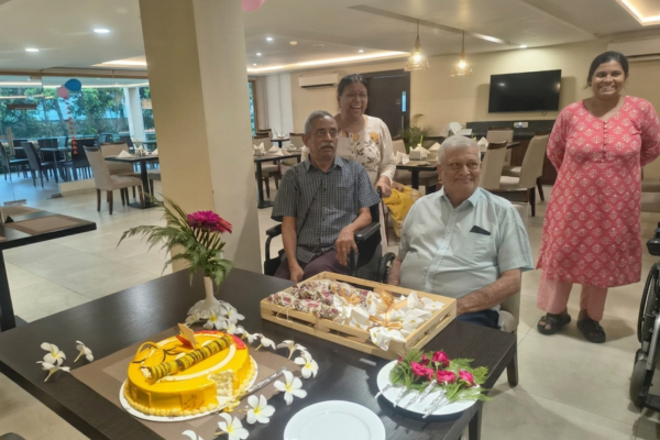 Senior residents smiling during a festive birthday celebration at a premium community recognized among the best senior living in India, featuring a decorated yellow cake and supportive care staff.