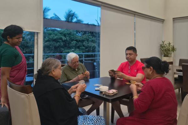 Senior citizens enjoying a card game together in a well-lit recreation lounge at a premier senior living in Kolkata community, assisted by a smiling staff member.
