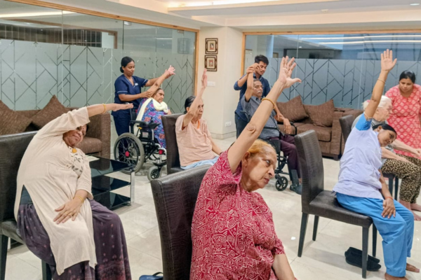 A group of senior citizens seated on dark leather chairs, performing side-stretch exercises with one arm raised overhead during a guided group session, with caregivers assisting in the background inside a modern indoor room.