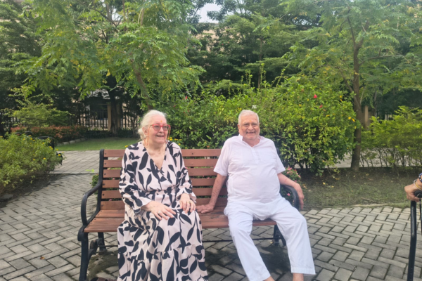 An elderly woman in a black-and-white patterned dress and an elderly man in a white outfit sitting together happily on a wooden park bench in a lush garden.