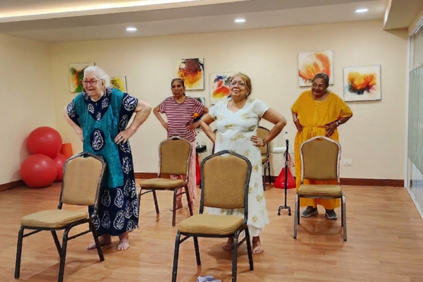 Senior residents performing standing physical therapy exercises using chairs for stability during a wellness session at a premium retirement facility.
