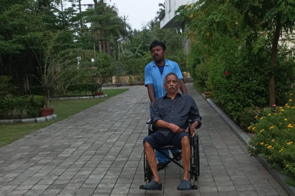 An elderly man in a wheelchair being assisted by a professional caregiver along a wide paved pathway inside a senior living community with lush green landscaping.