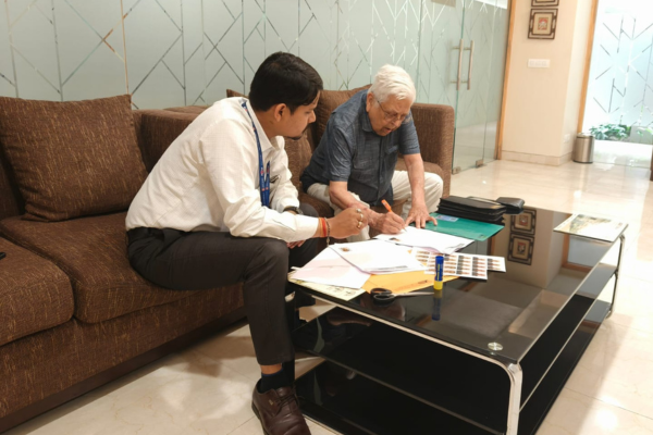 An advisor is assisting an elderly man with paperwork at a glass table, highlighting premium senior citizen care Kolkata services inside a comfortable, modern residential lounge.