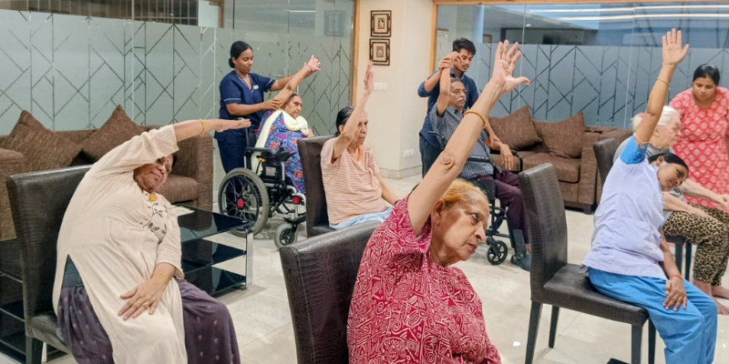 enior citizens practicing chair yoga and stretching exercises guided by supportive caregivers in a spacious recreation room at a premier facility offering luxury senior living in India.