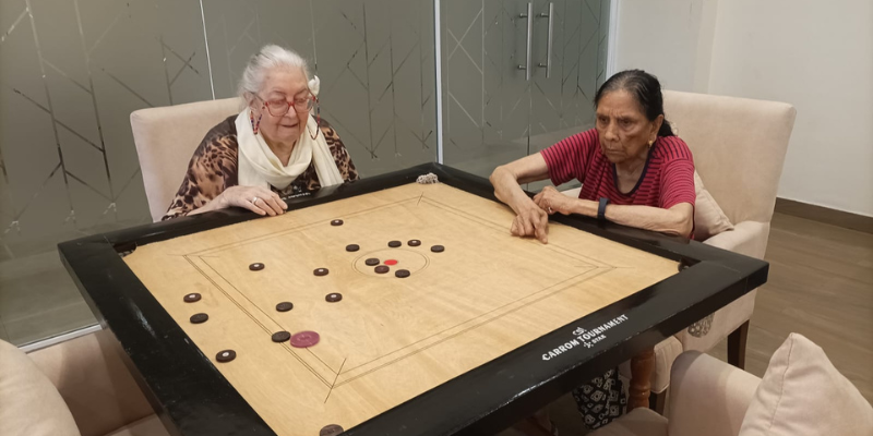 Two elderly women playing a competitive game of carrom on a large wooden board while seated in comfortable armchairs inside a modern senior community recreation room.