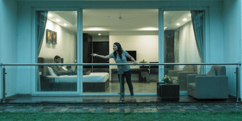 A woman reaching out to feel raindrops on a spacious private balcony while a man works on a laptop inside a modern suite during a monsoon staycation.