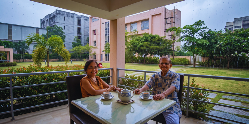 Senior couple enjoying tea on a covered balcony overlooking green lawns during a peaceful rainy day at a luxury senior living wellness retreat.
