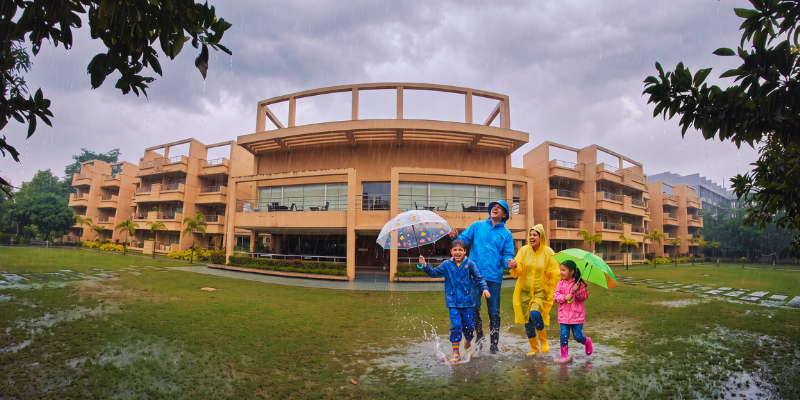 A happy family wearing colourful raincoats and holding umbrellas splashing in rain puddles on a resort lawn during a monsoon vacation.