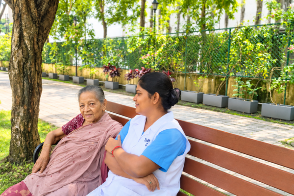 An elderly woman in traditional Indian attire sitting on a wooden park bench alongside a smiling carer in a white uniform.